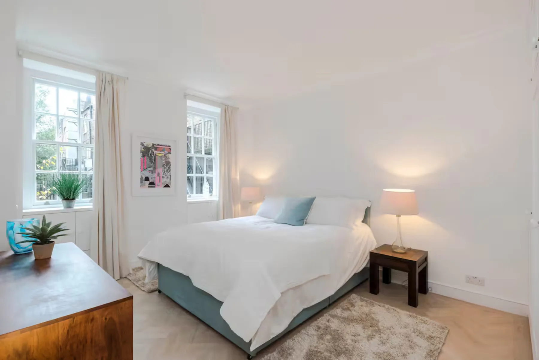 A bright bedroom with a white bed, two windows, and a bedside table.