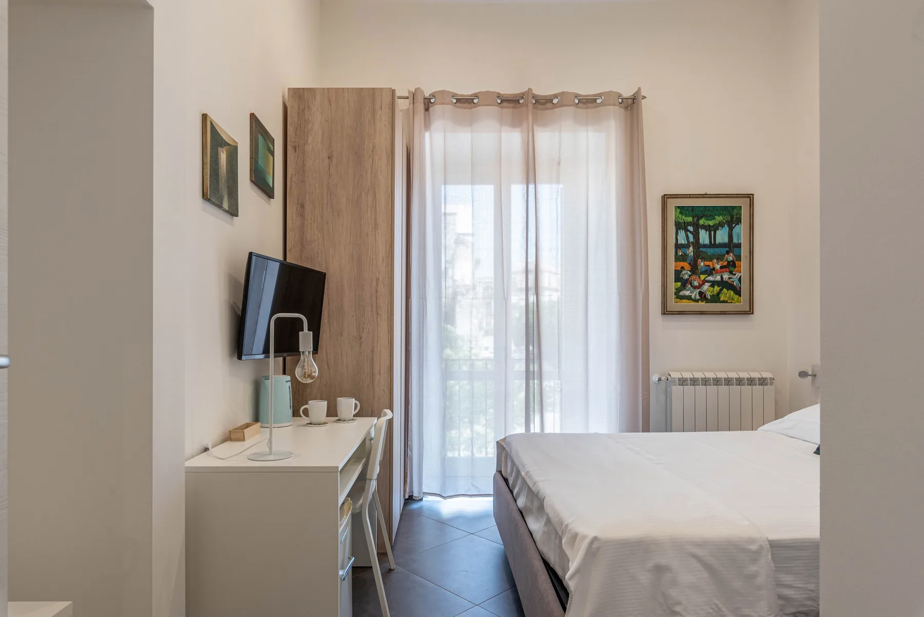 A bedroom with a bed, desk, and sheer curtains over the window.