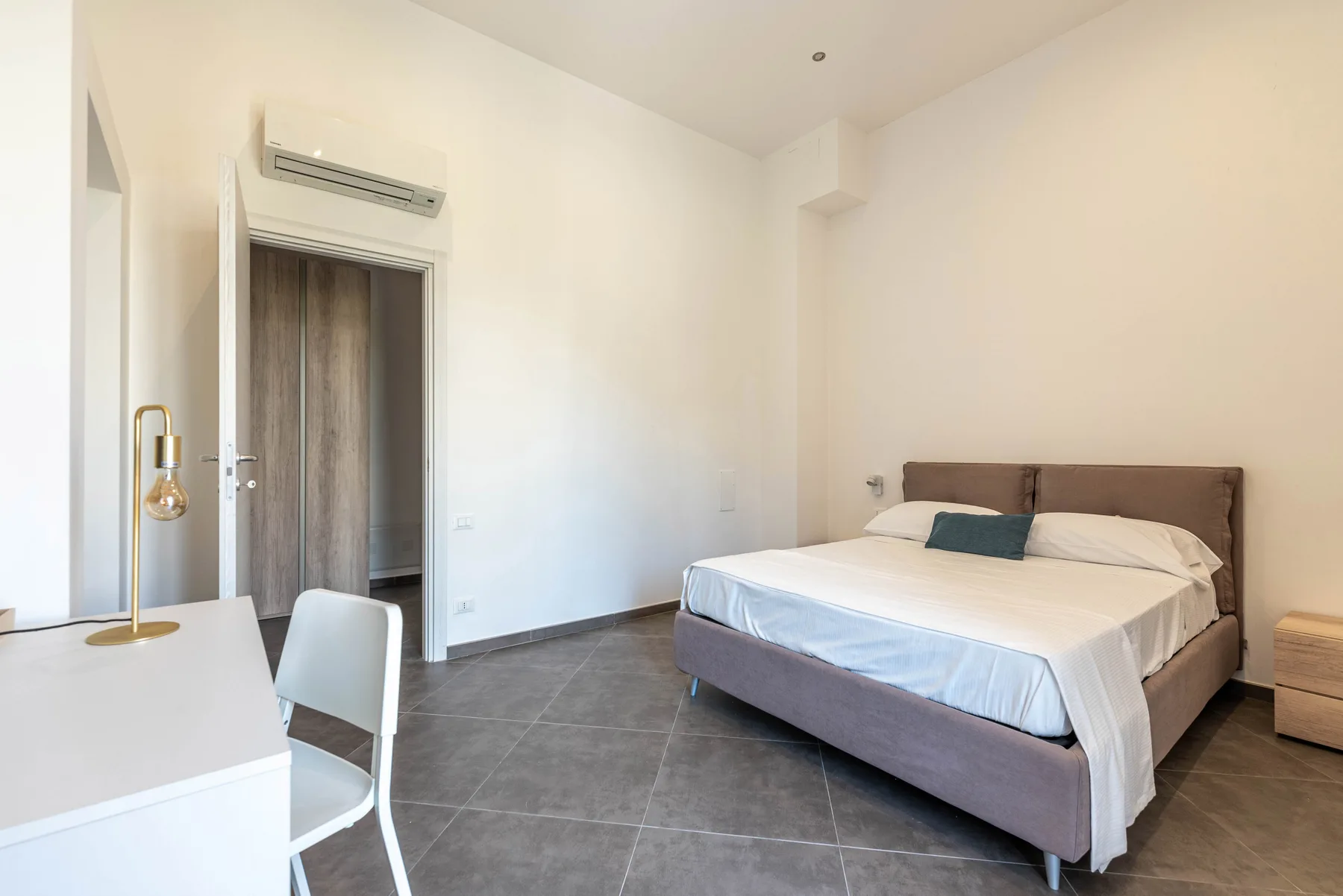 A bright bedroom with a double bed, bedside table, and wall-mounted air conditioner.