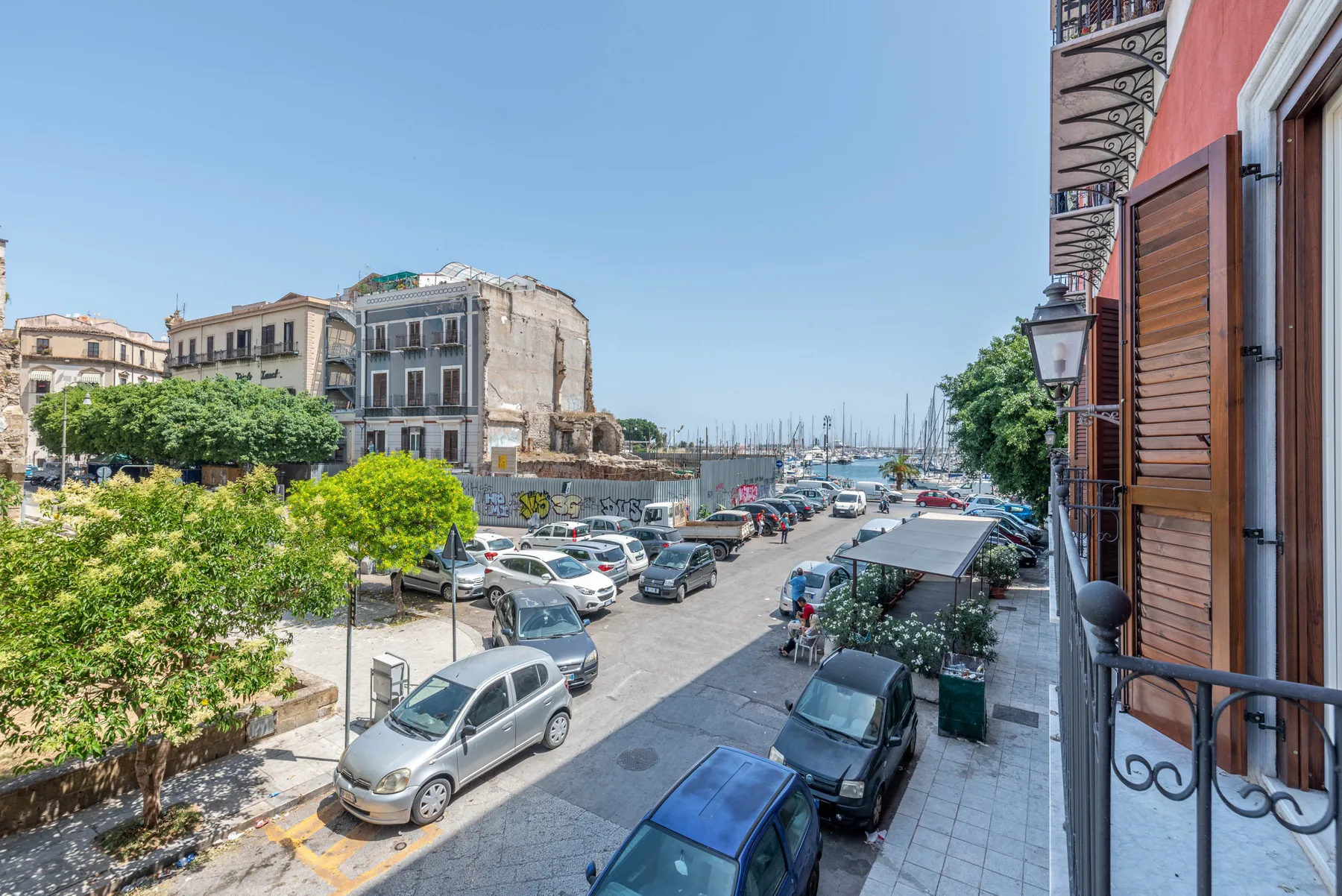 A sunny street with parked cars leads toward a marina and boats.