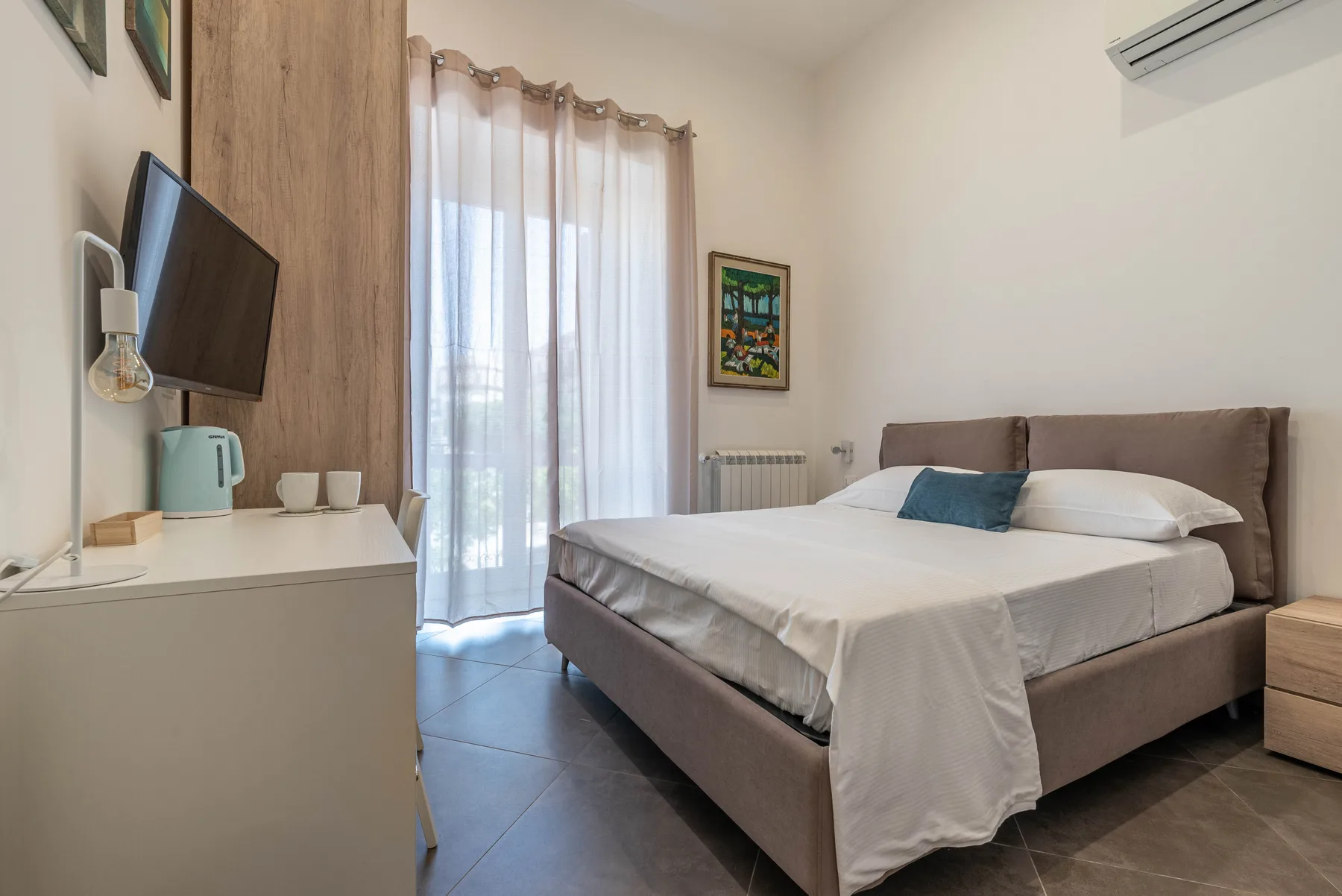 A bedroom with a neatly made bed, sheer curtains, and a television.