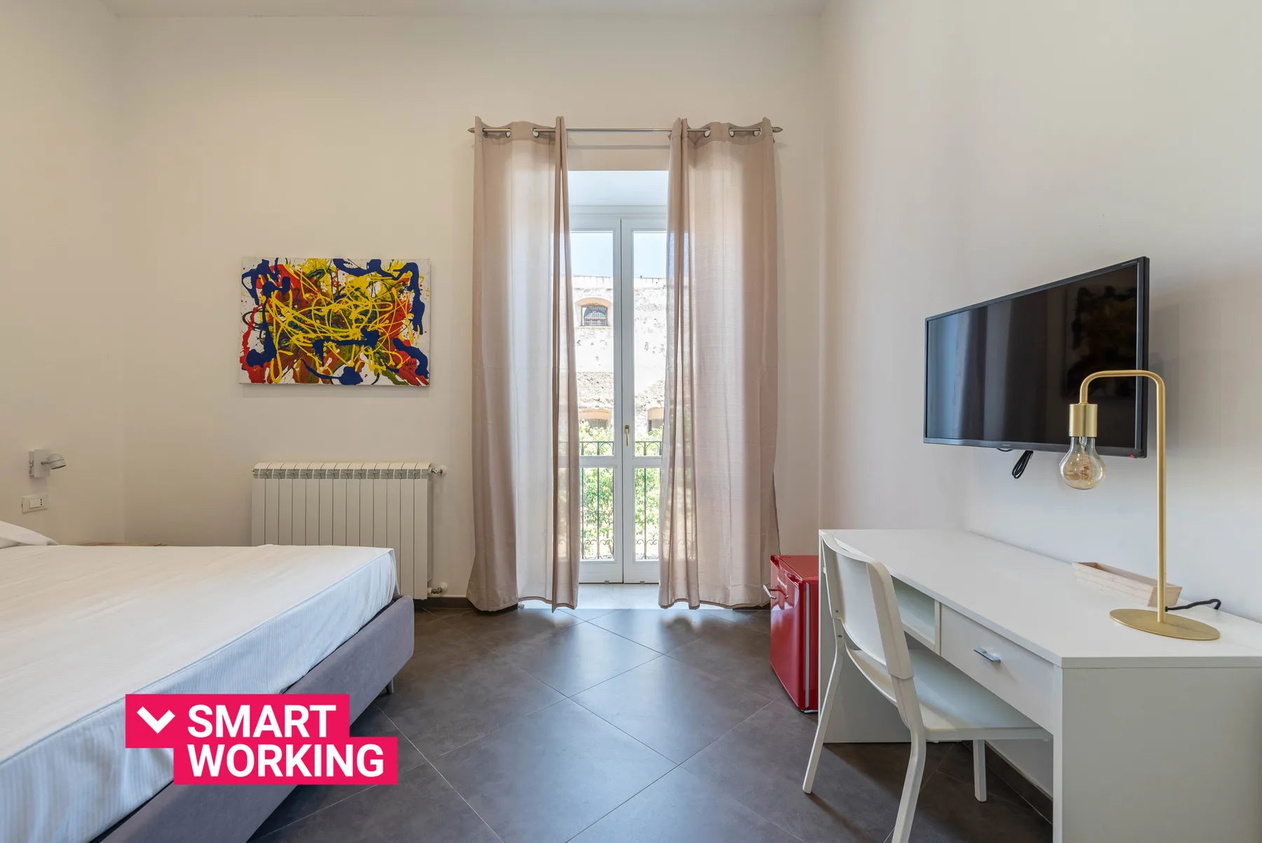 A bright bedroom with a bed, desk, and glass doors opening onto a balcony.