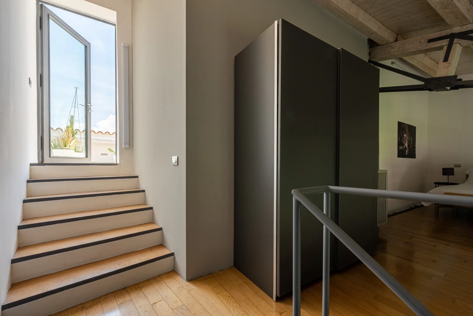 A bright loft interior with stairs beside a tall dark partition and a bed in the background.