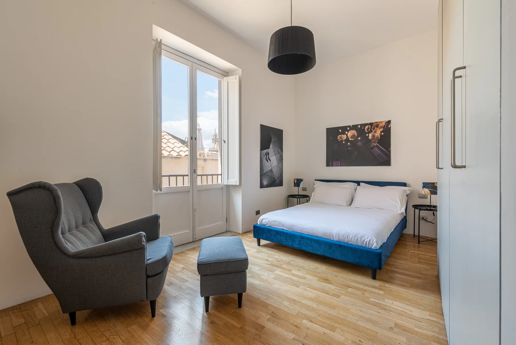 A bedroom with a bed, an armchair, and a window overlooking nearby rooftops.