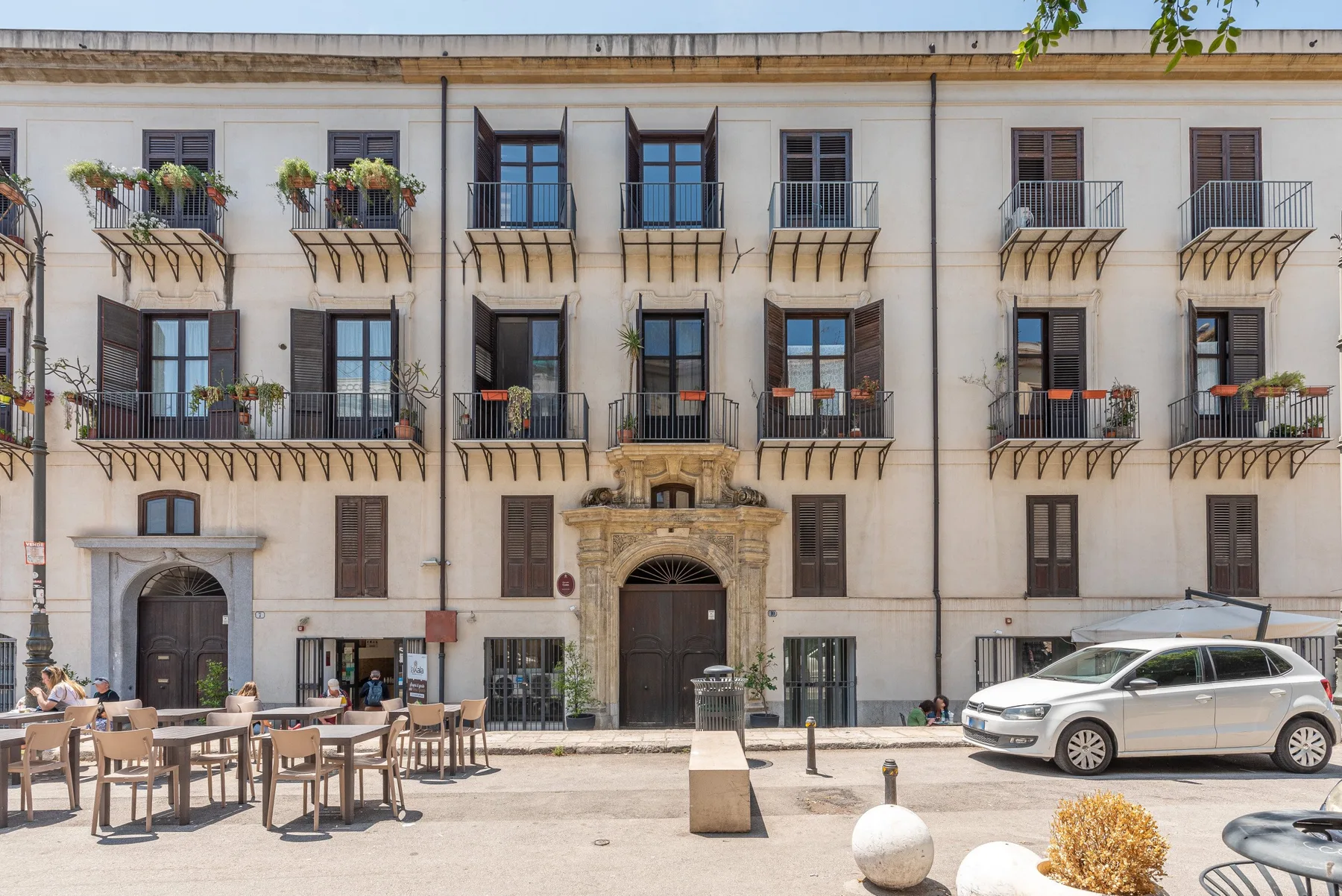 A multi-story building with balconies faces an outdoor seating area and parked cars.