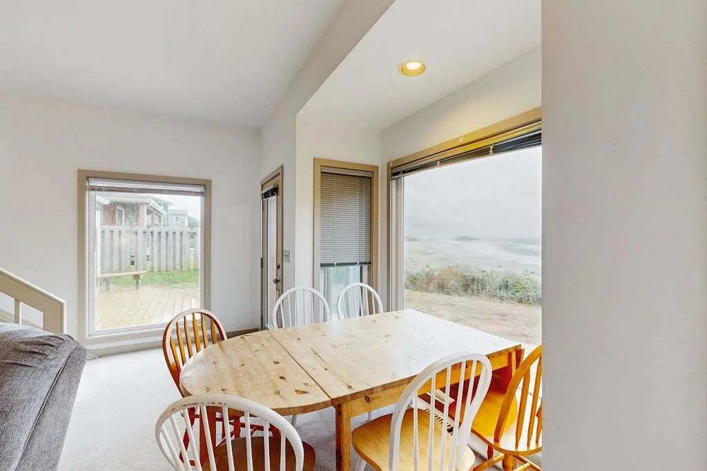 Una luminosa zona de comedor tiene una mesa de madera junto a grandes ventanas con vistas al agua.