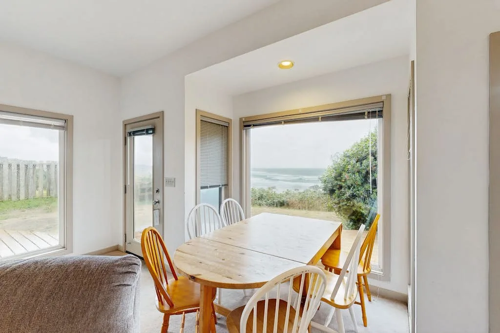 Un comedor luminoso con una mesa de madera junto a grandes ventanas con vistas al agua.