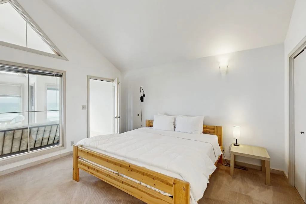 Un dormitorio luminoso con cama de madera, ropa de cama blanca y grandes ventanas.