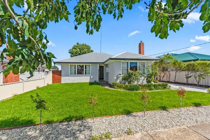 A single-story house faces a green lawn and gravel path beneath leafy branches.