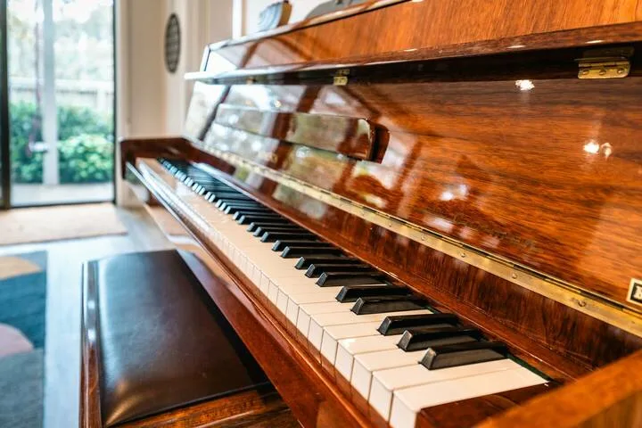 An upright piano stands beside a large window in a bright room.