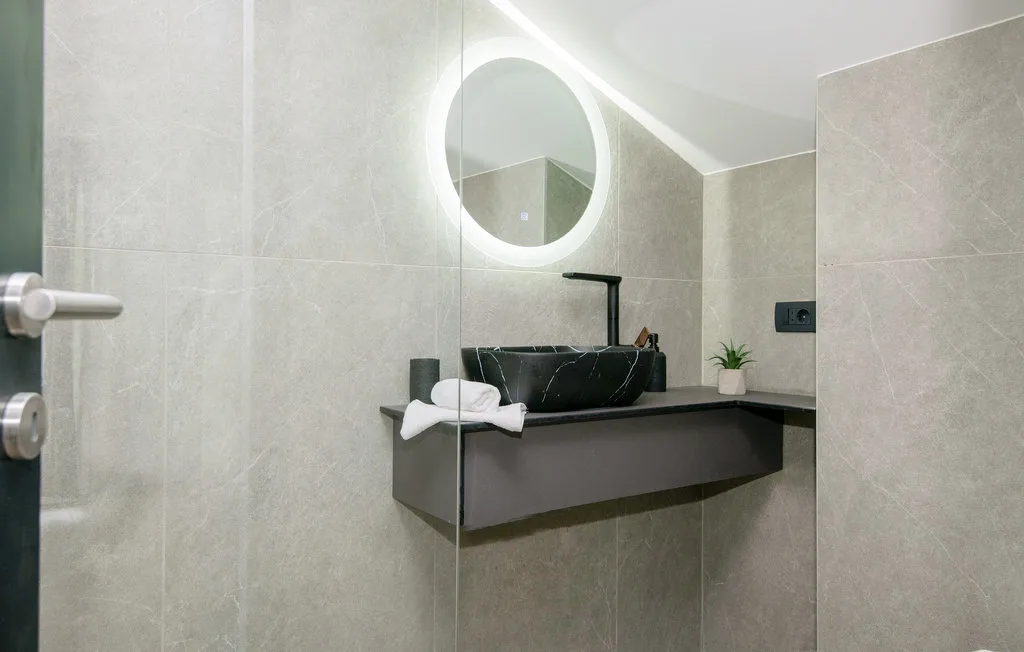 Un tocador de baño moderno con espejo redondo iluminado y lavabo oscuro.