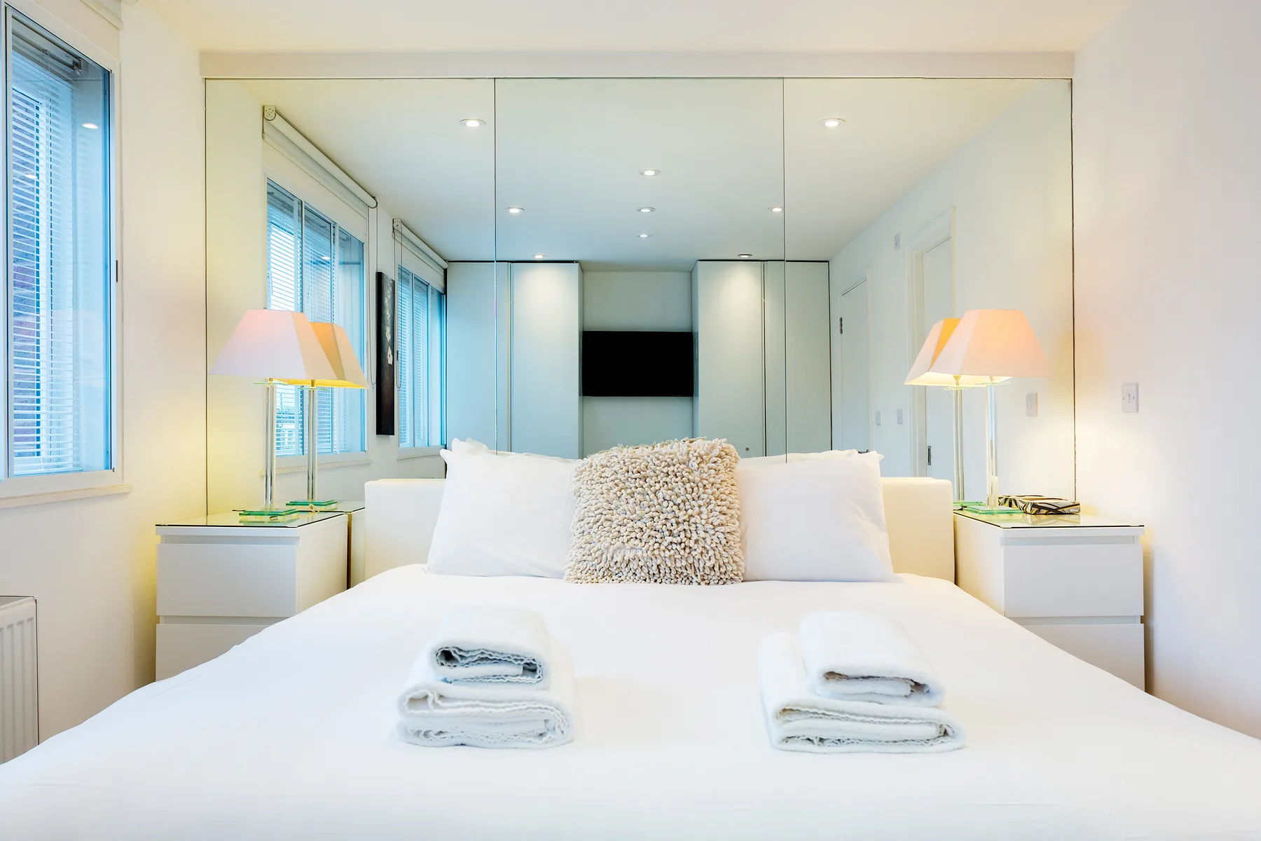Un dormitorio luminoso con una cama grande, paredes espejadas y ventanas a un lado.
