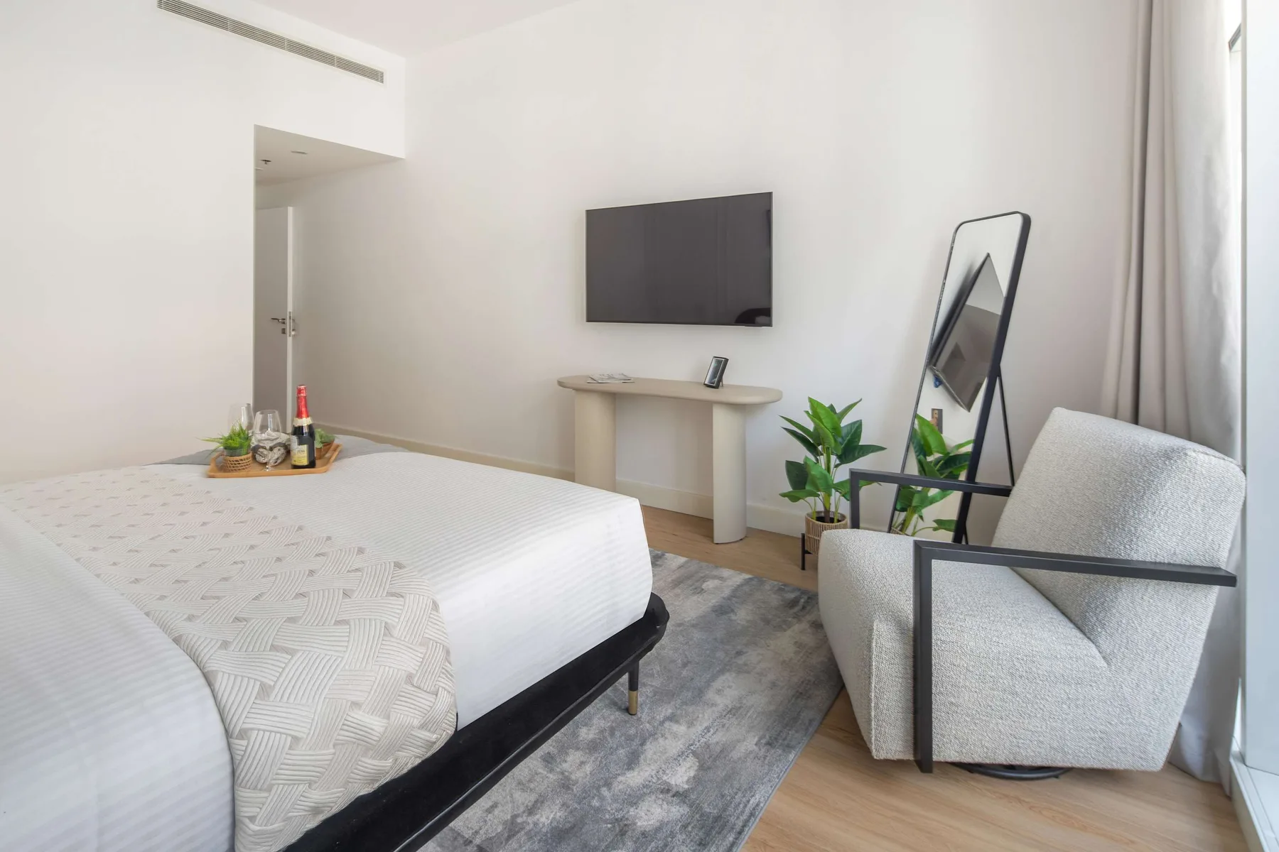 Un dormitorio luminoso con cama, televisor en la pared y una zona de descanso junto a la ventana.