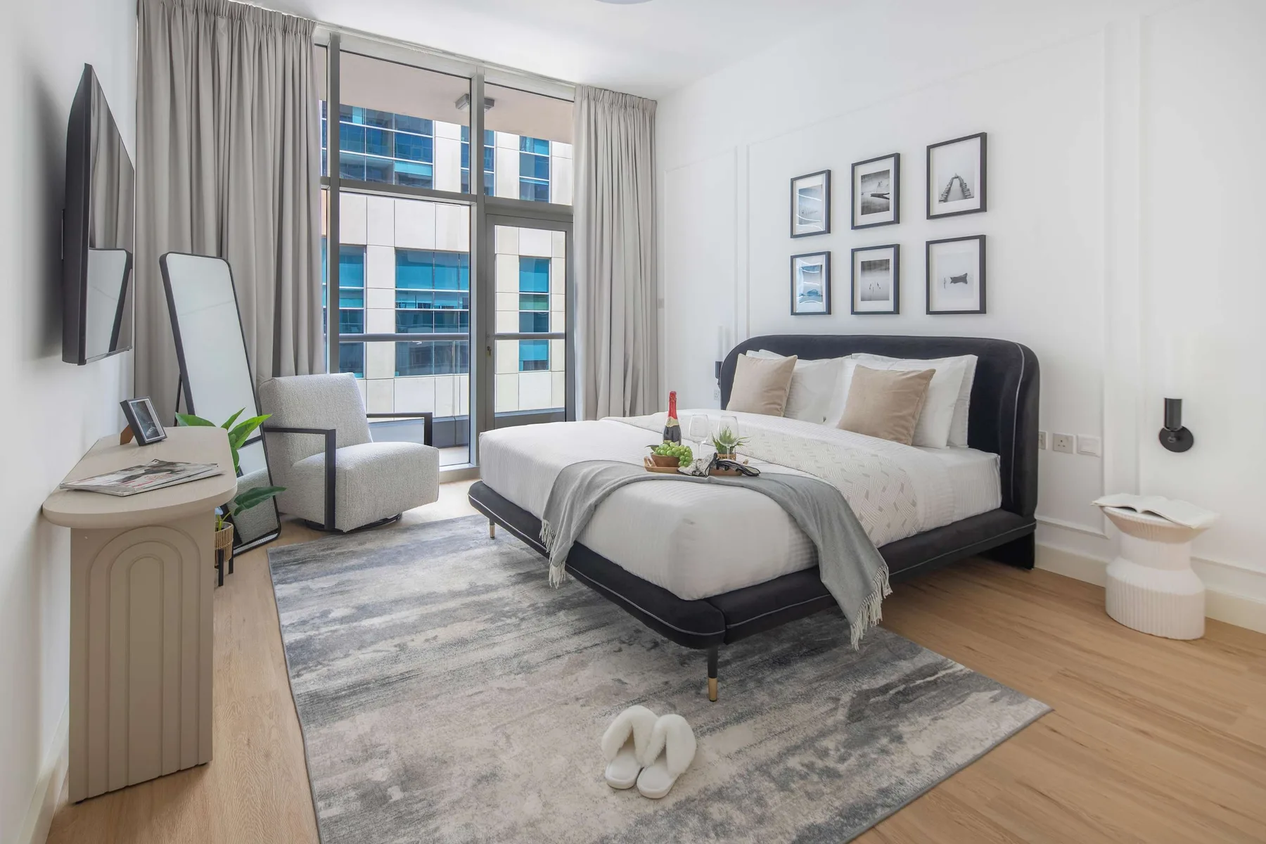 Un dormitorio luminoso con una cama, mesitas de noche y ventanas con vistas a edificios cercanos.