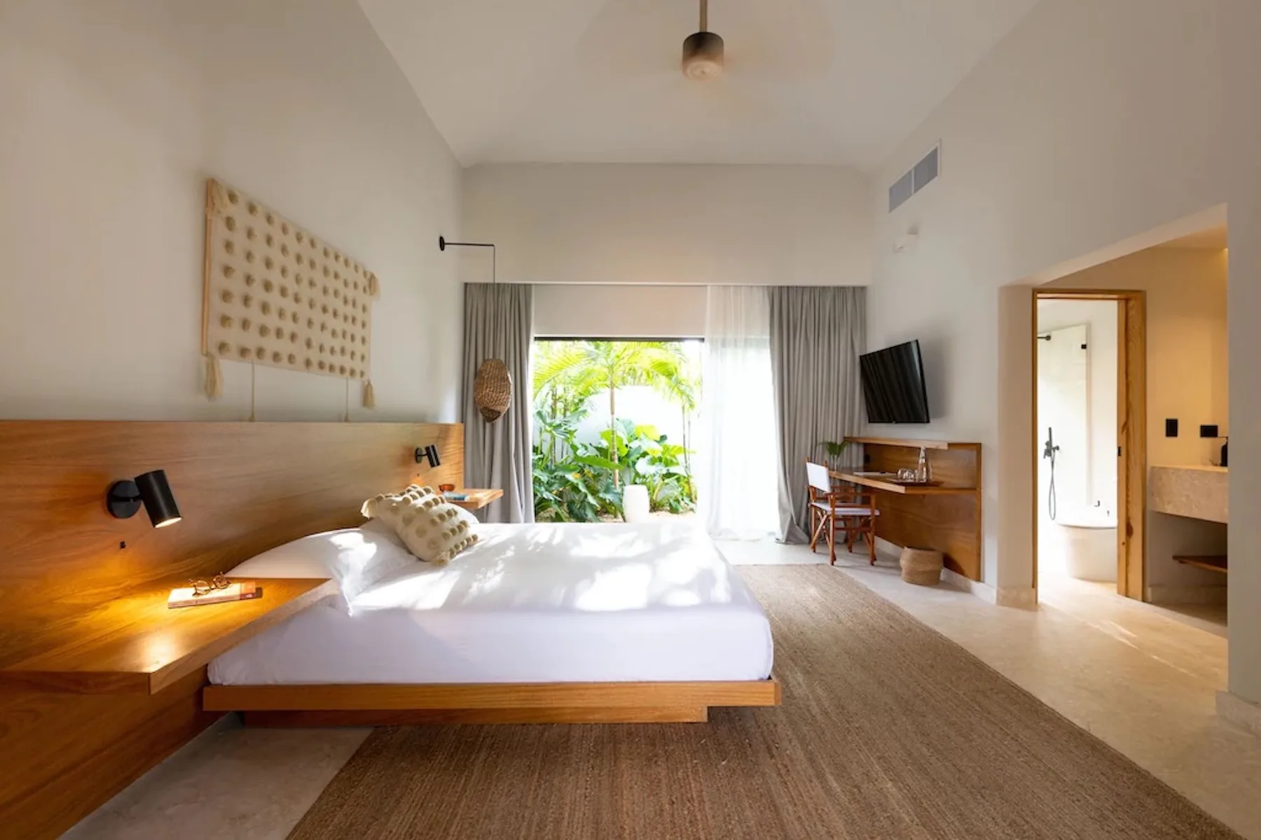 A bright bedroom with a bed, wooden bedside surfaces, and a glass door opening to greenery.