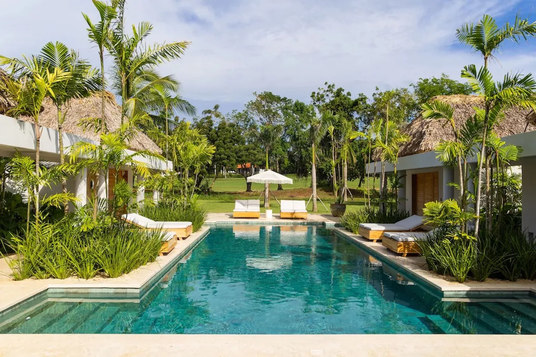 A swimming pool sits between tropical greenery and thatched-roof buildings.