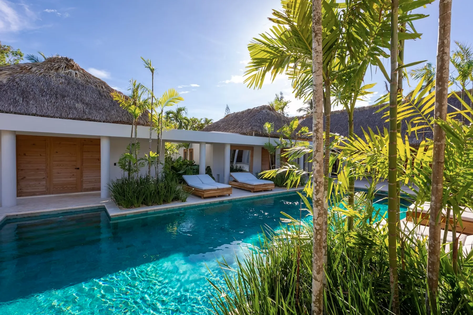 A tropical pool beside a low building with shaded lounge seating.