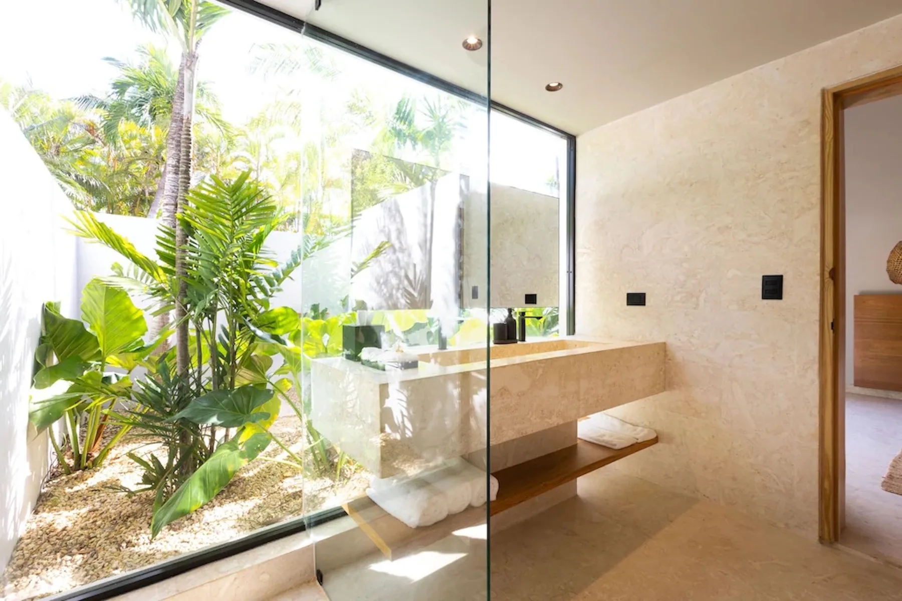 A bathroom vanity sits beside a floor-to-ceiling glass wall overlooking tropical plants.