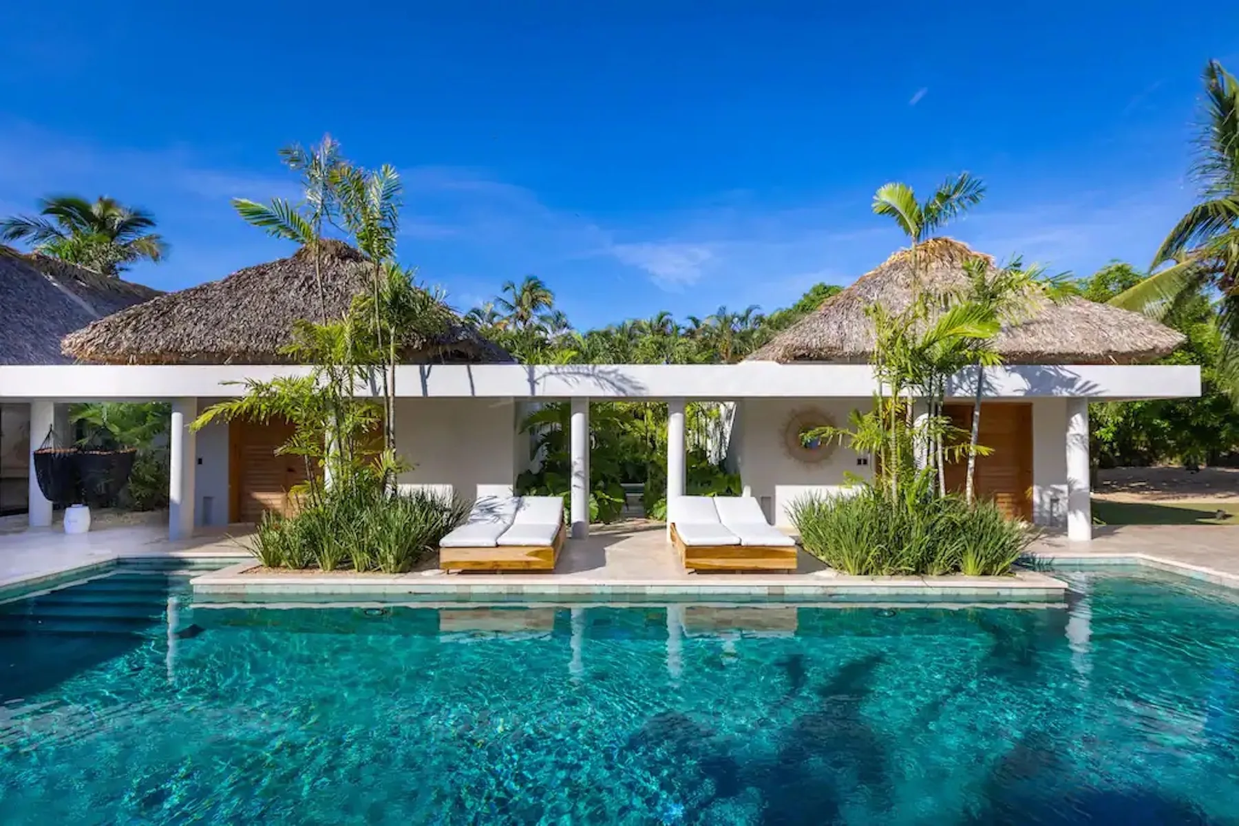 A tropical resort-style pool fronts a low building with thatched roofs and greenery.