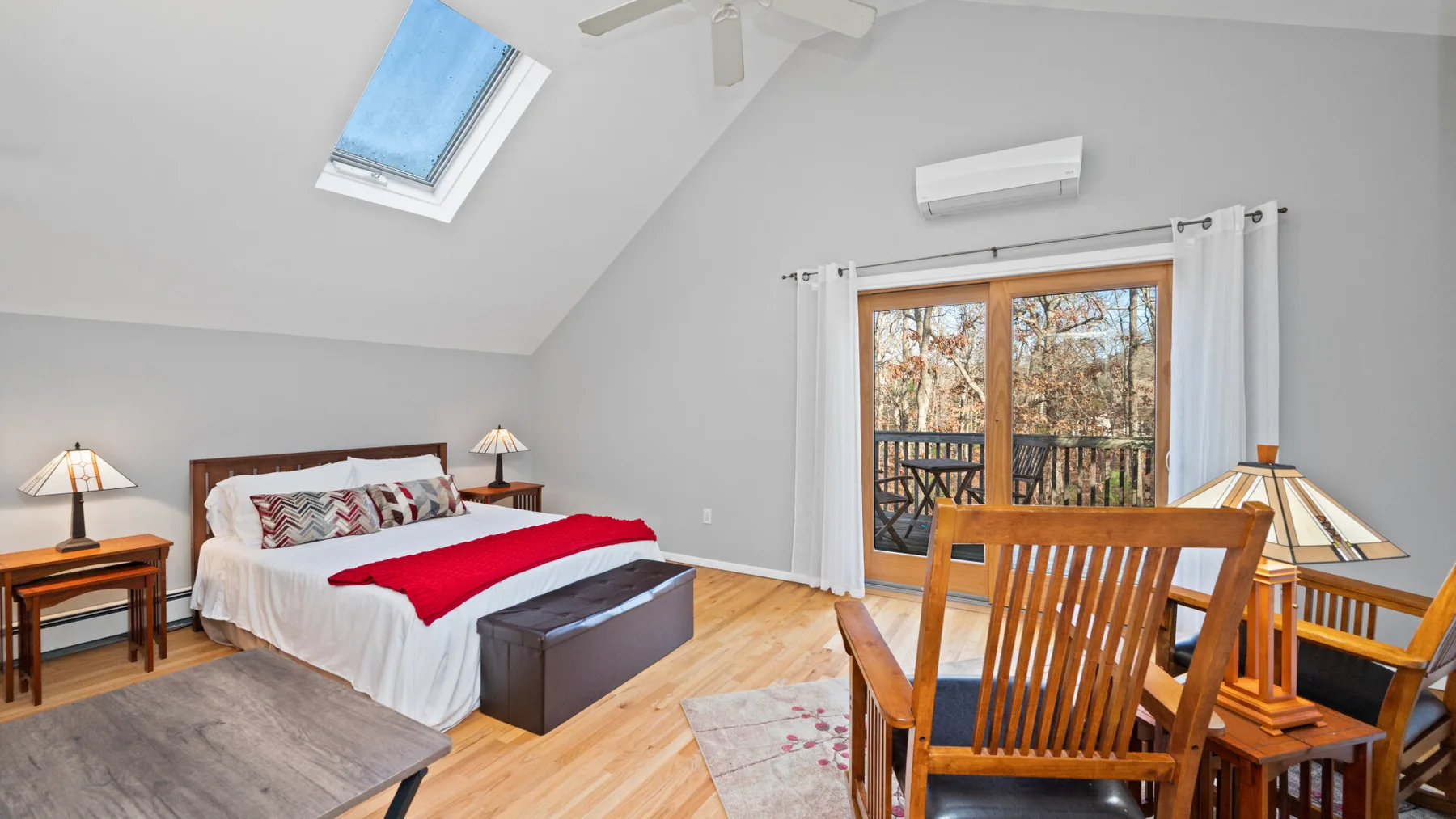 A bright bedroom with a large bed, wooden seating, and glass doors opening to a balcony.
