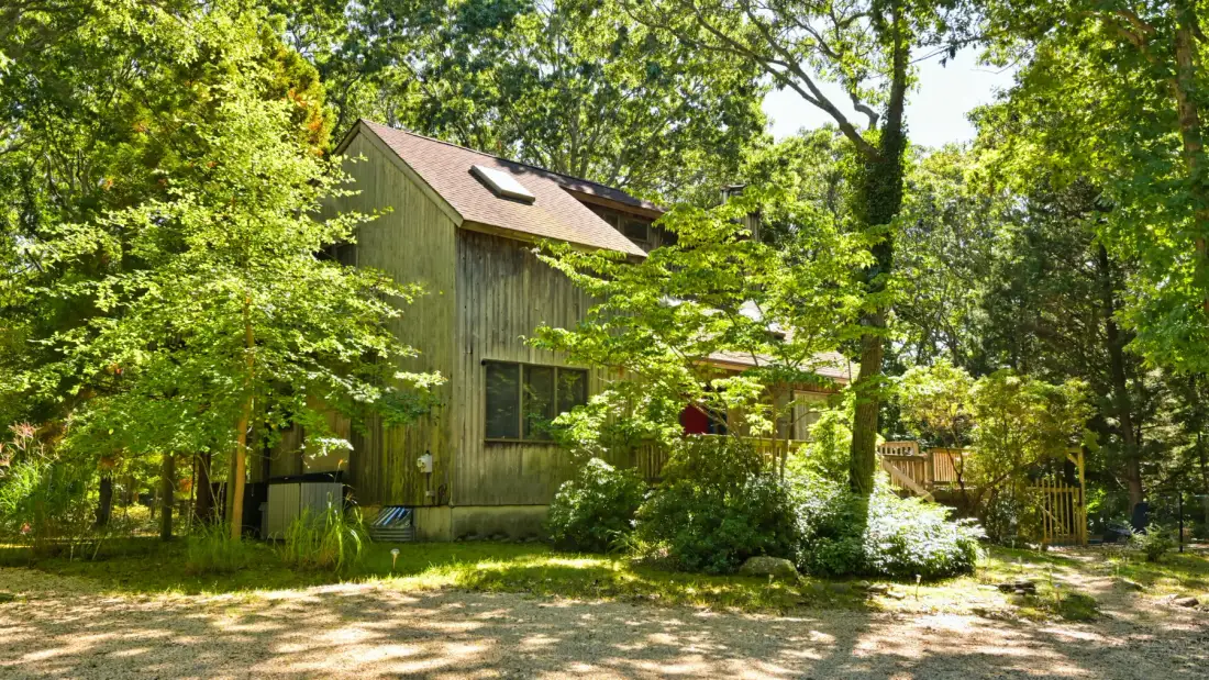A wooded house stands among leafy trees beside a sunlit gravel drive.