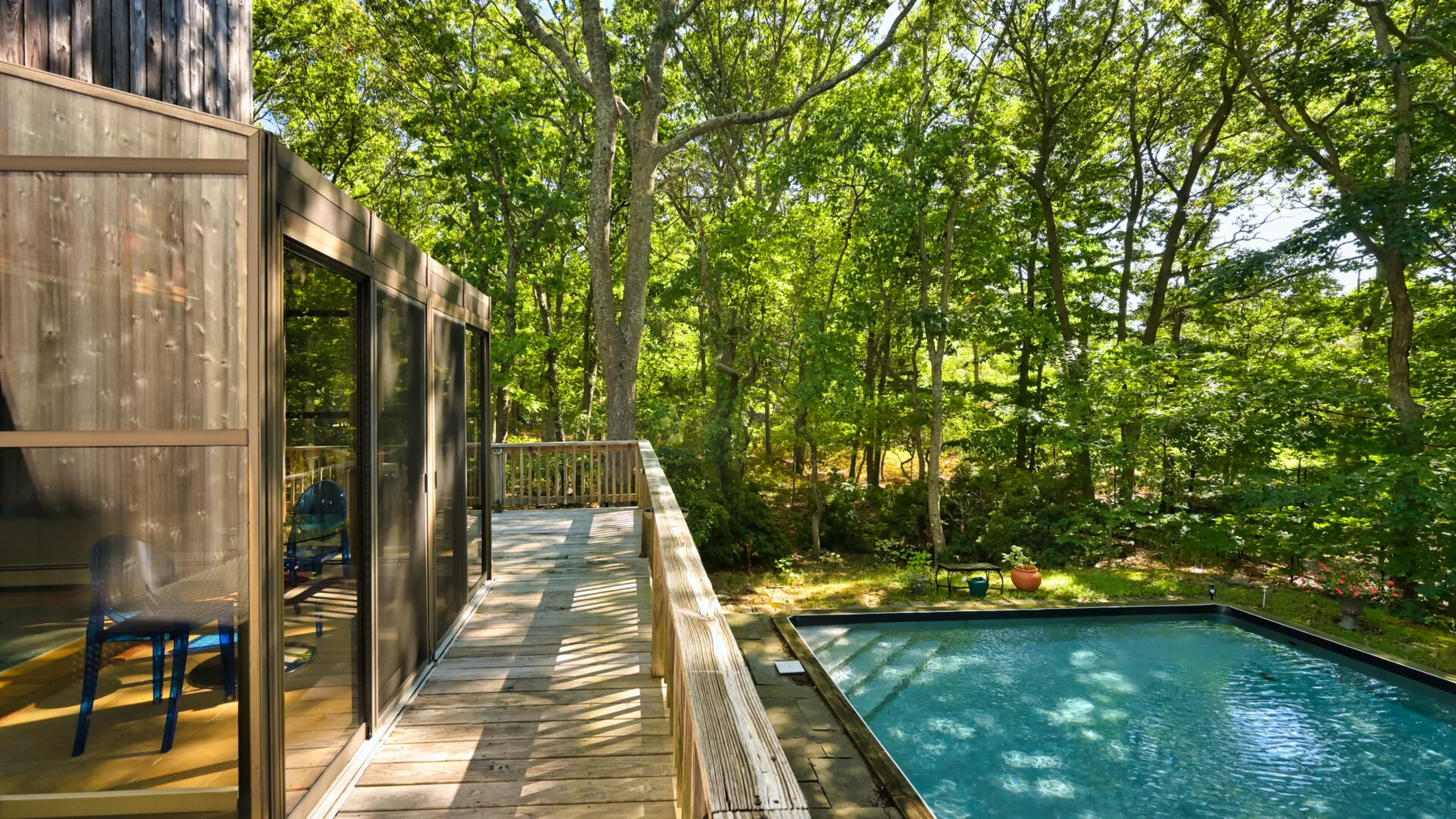 A wooden deck beside a swimming pool, surrounded by leafy trees.