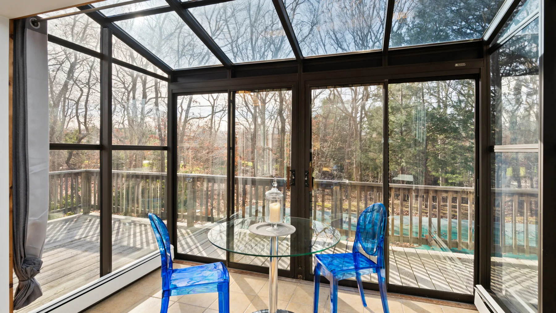 A glass-enclosed sunroom with a round table and blue chairs overlooks trees.