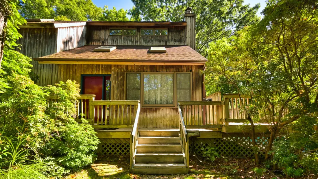 A wooden cabin surrounded by trees, with a raised deck and front steps.