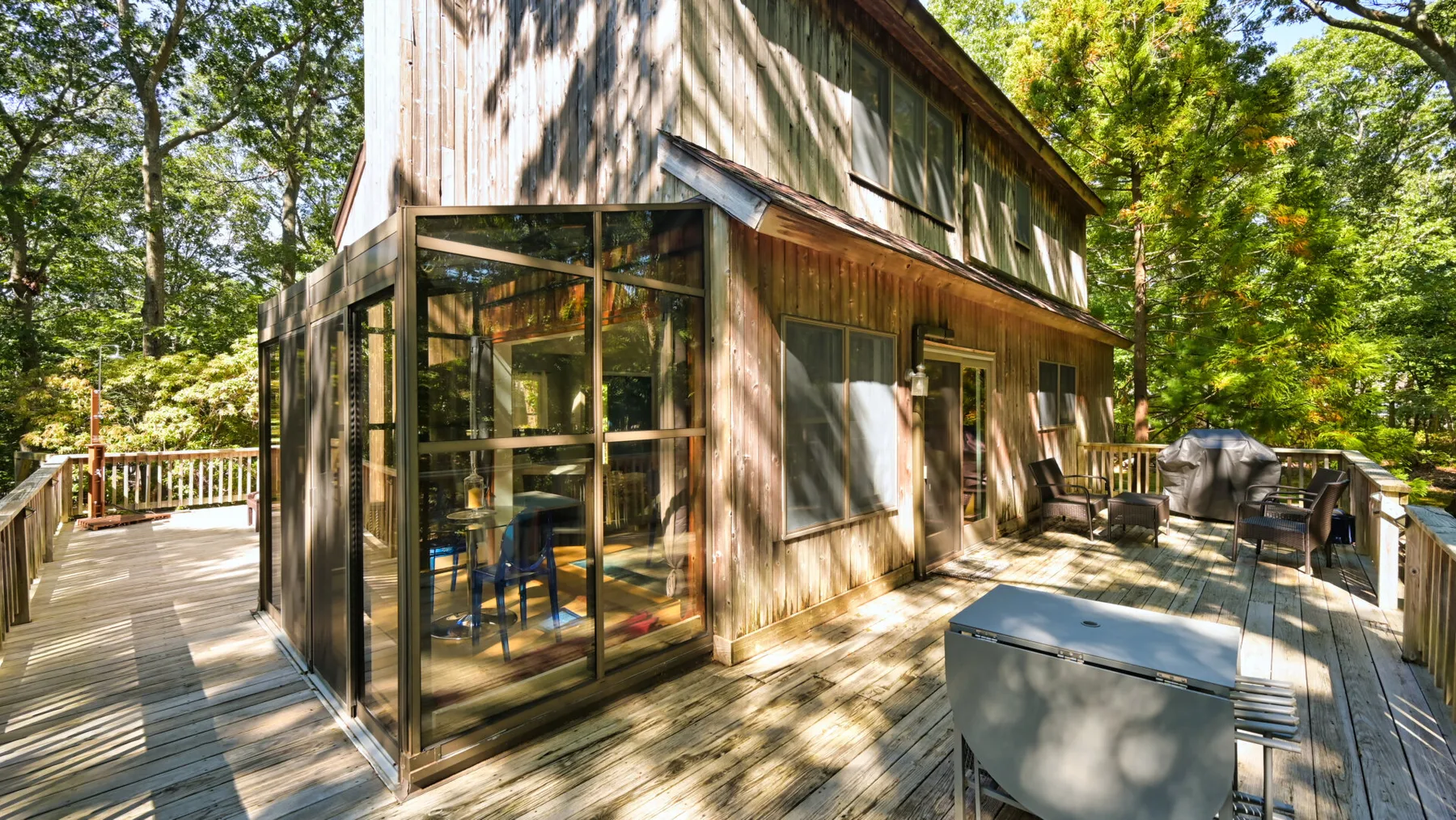 A wooden house opens onto a broad outdoor deck surrounded by trees.