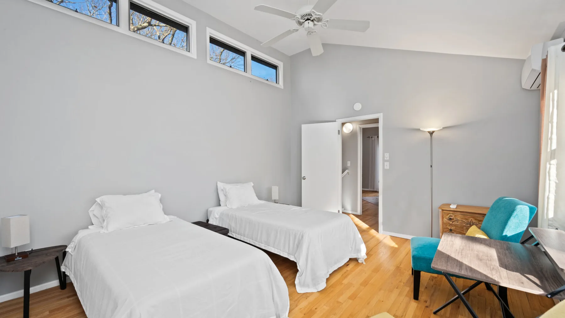 A bright bedroom with two beds, a desk, and high windows.