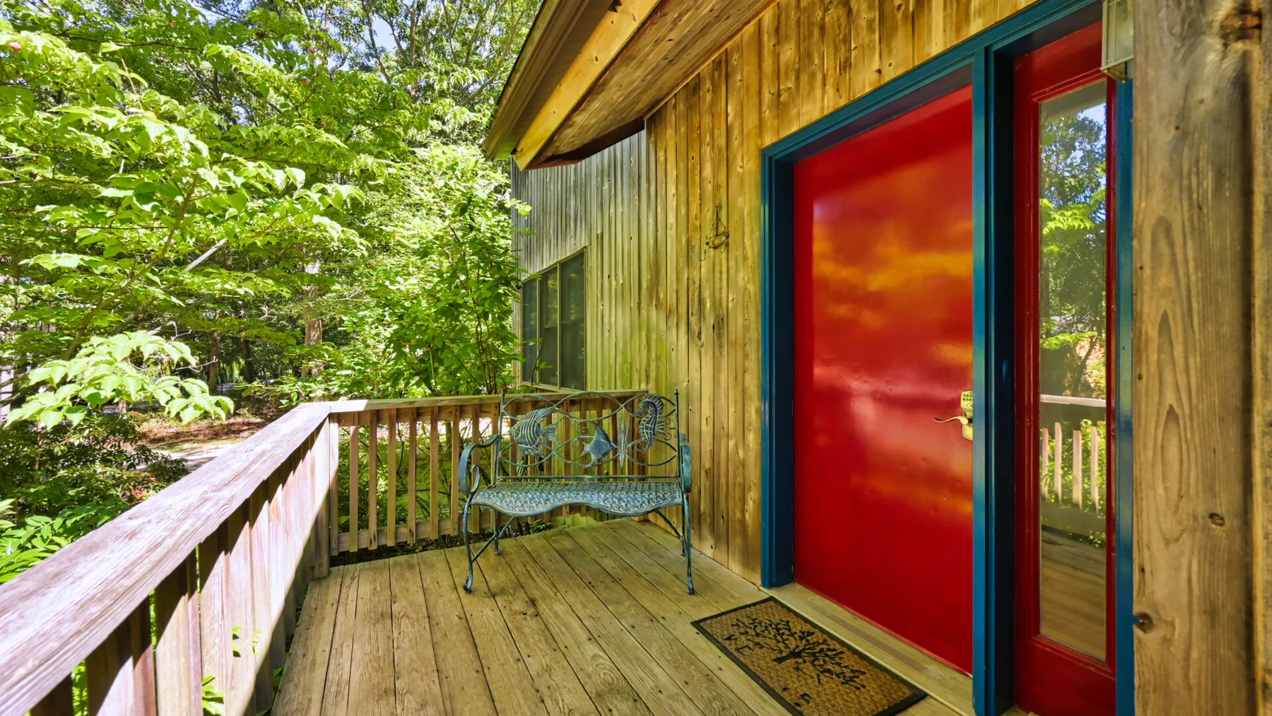 A wooden porch with a bright red door and a metal bench overlooks leafy trees.
