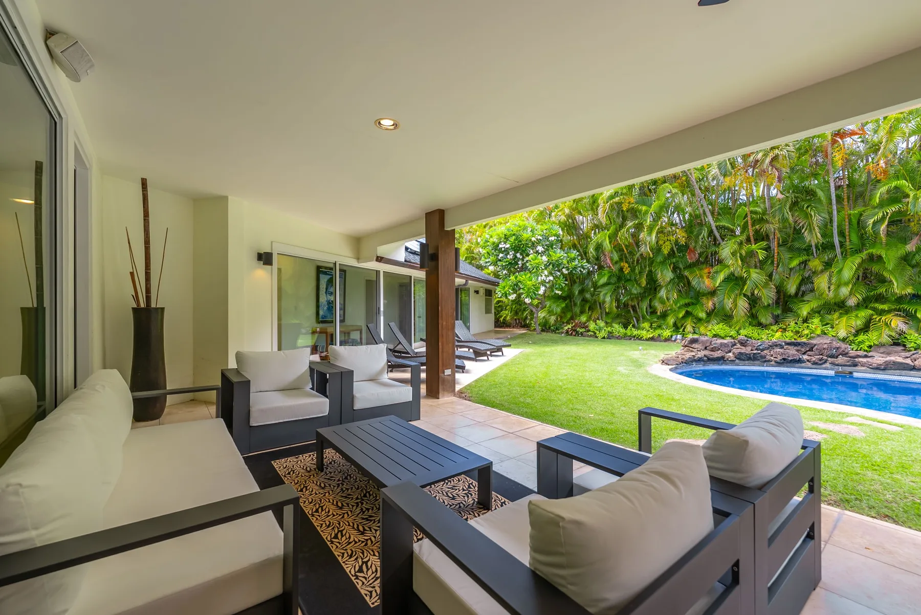 Una zona de estar cubierta da a un césped, una piscina y vegetación tropical.
