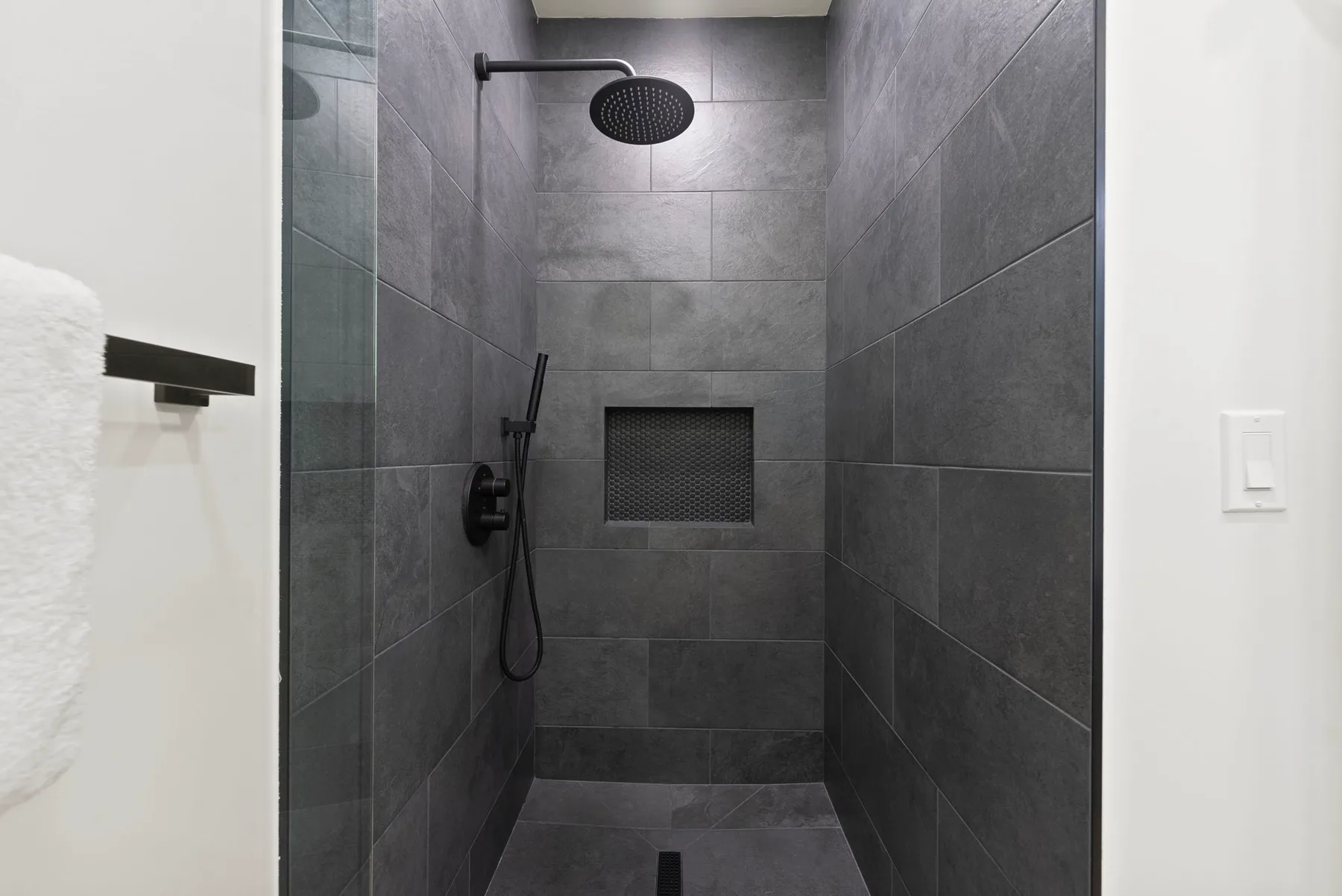 Una ducha de azulejos oscuros con rociador superior redondo y nicho en la pared.