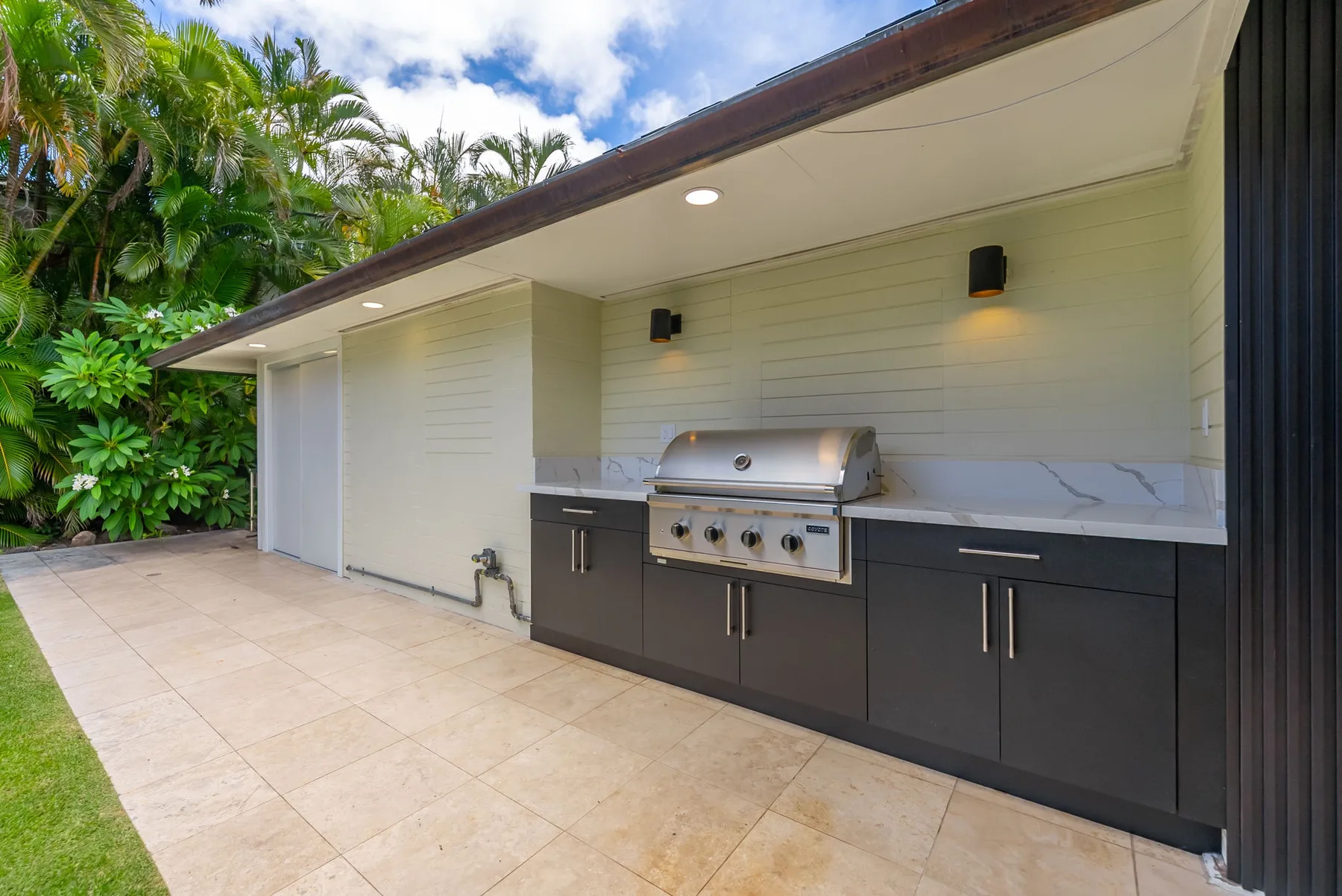 Una cocina exterior con parrilla empotrada y armarios oscuros está junto a vegetación tropical.