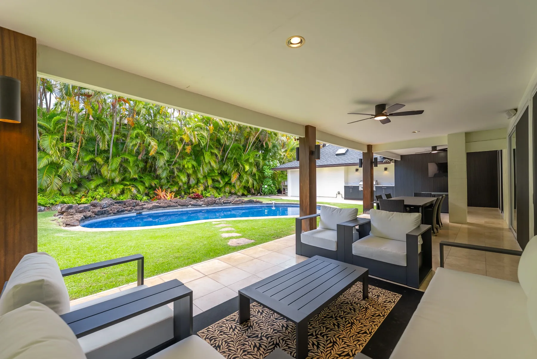 Un patio cubierto con asientos exteriores da a una piscina y vegetación tropical.