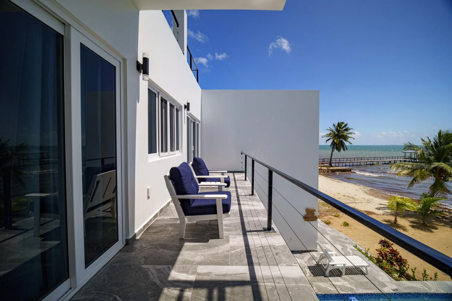 A sunny balcony with outdoor seating overlooks the beach and ocean.