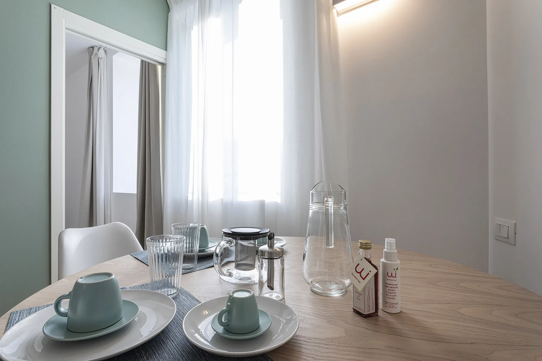 Una mesa con tazas y vasos está junto a una ventana luminosa con cortinas.