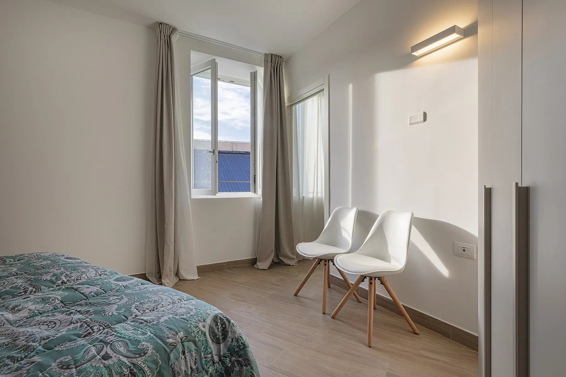 Un dormitorio luminoso con dos asientos blancos junto a una ventana alta.