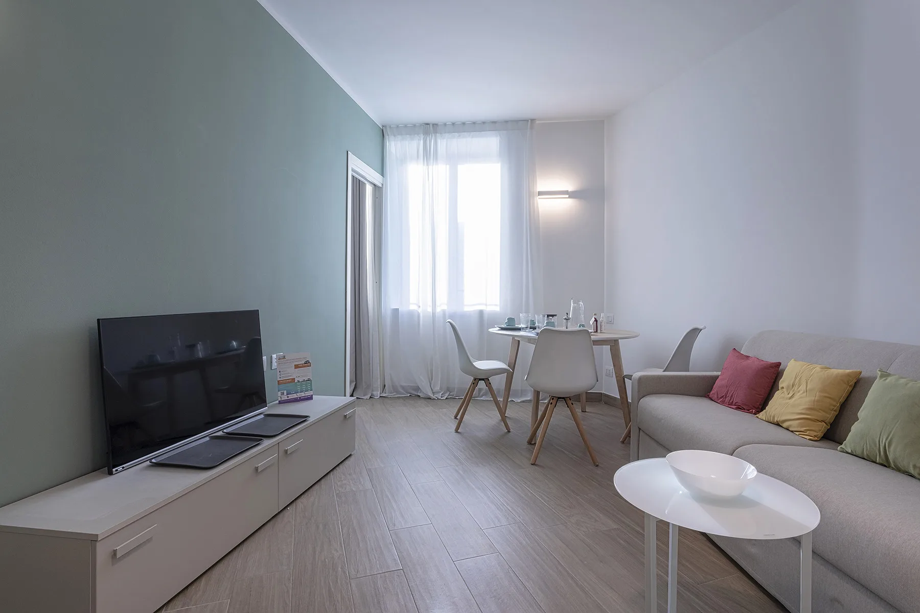 Una sala luminosa con asientos, un televisor y una pequeña mesa de comedor junto a la ventana.