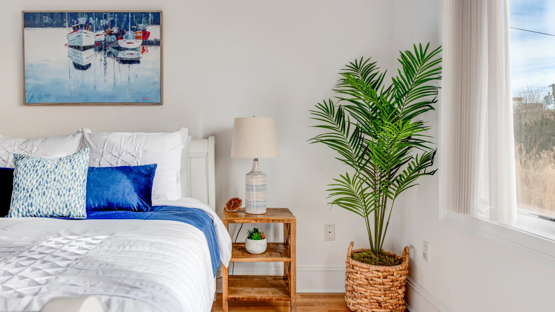 A bright bedroom with a bed, bedside table, and potted palm beside a window.