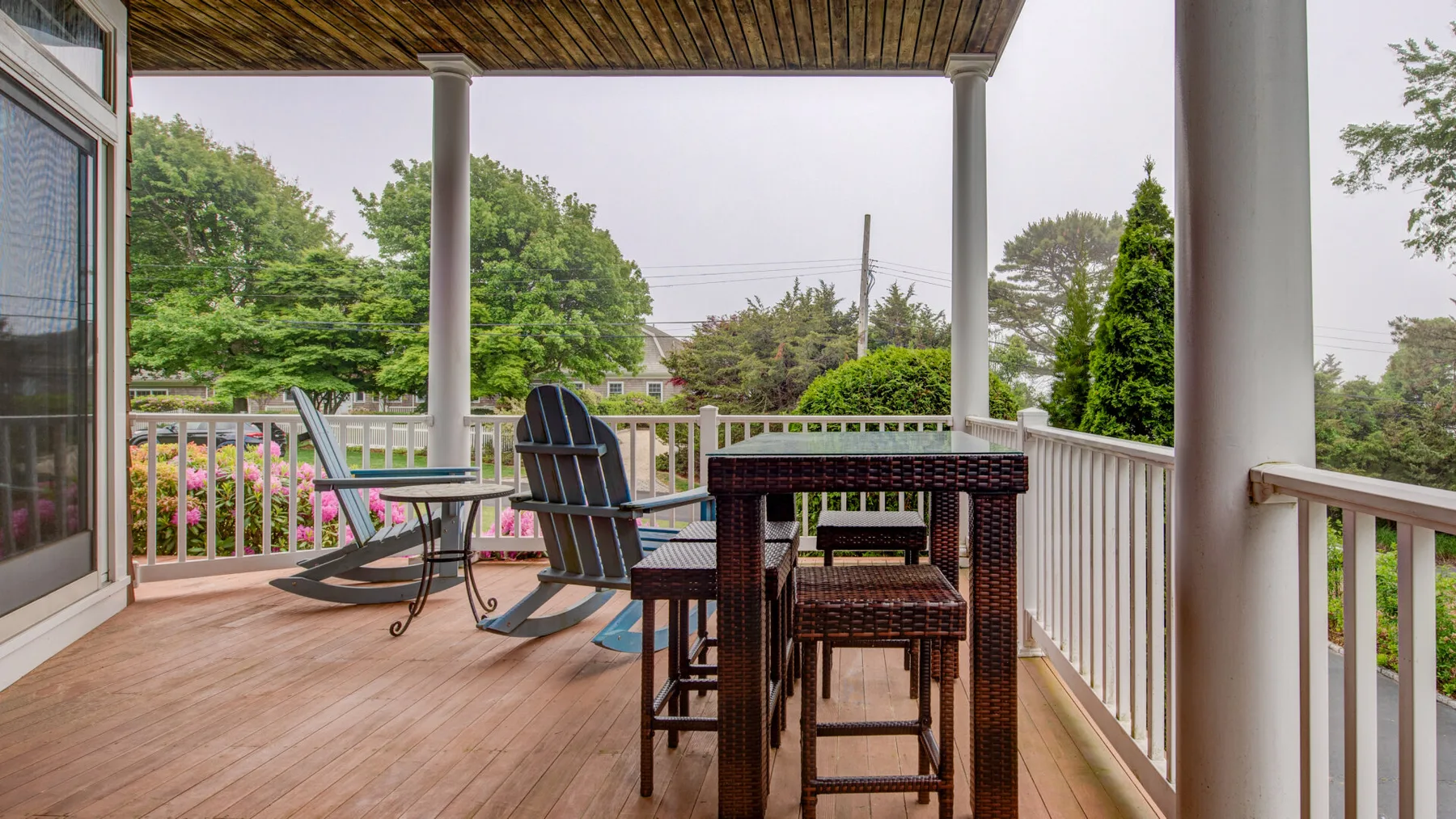 A covered outdoor deck with dining furniture and seating overlooks leafy trees.