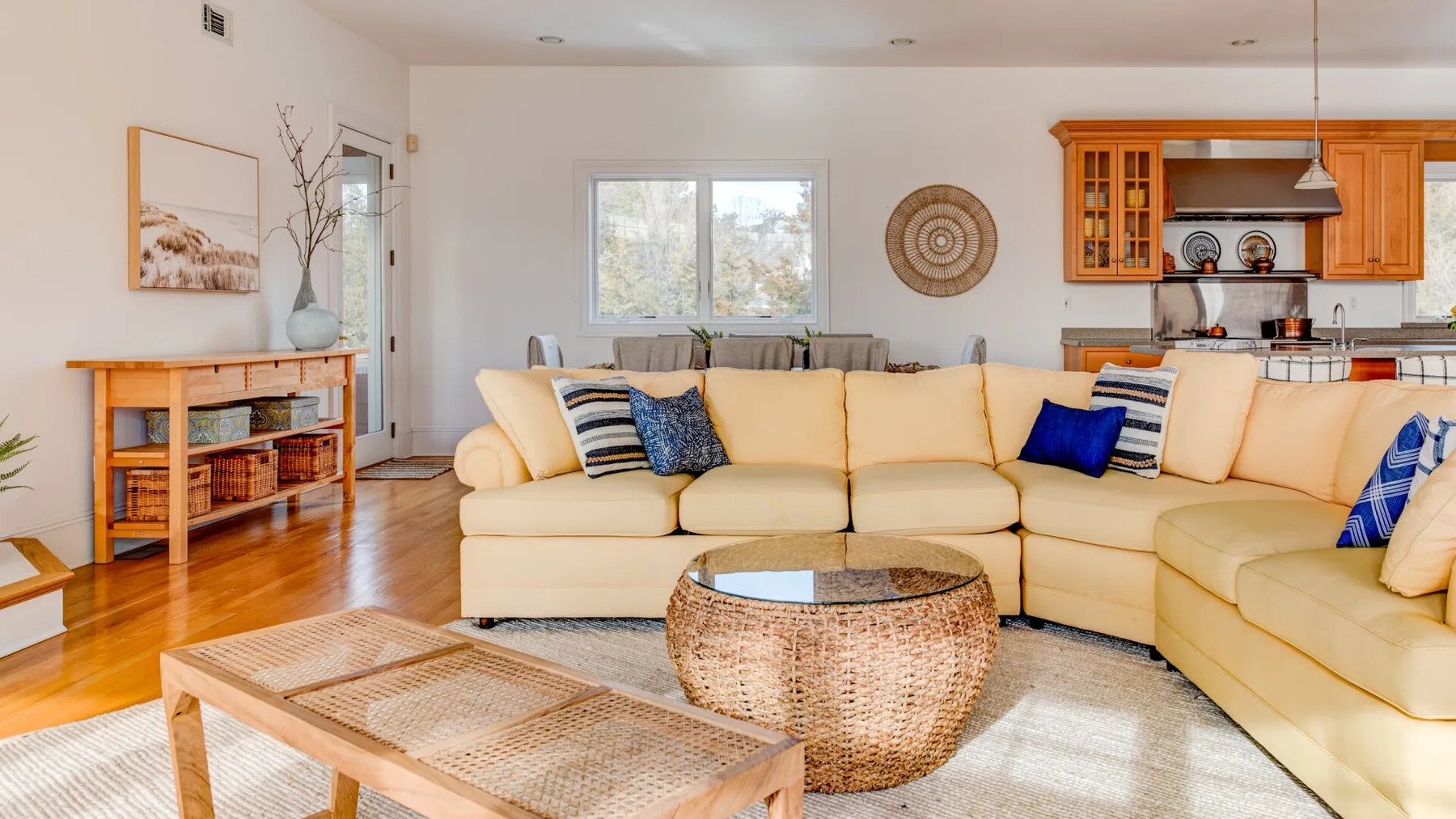 A bright living room with cream seating, a woven coffee table, and an open kitchen.