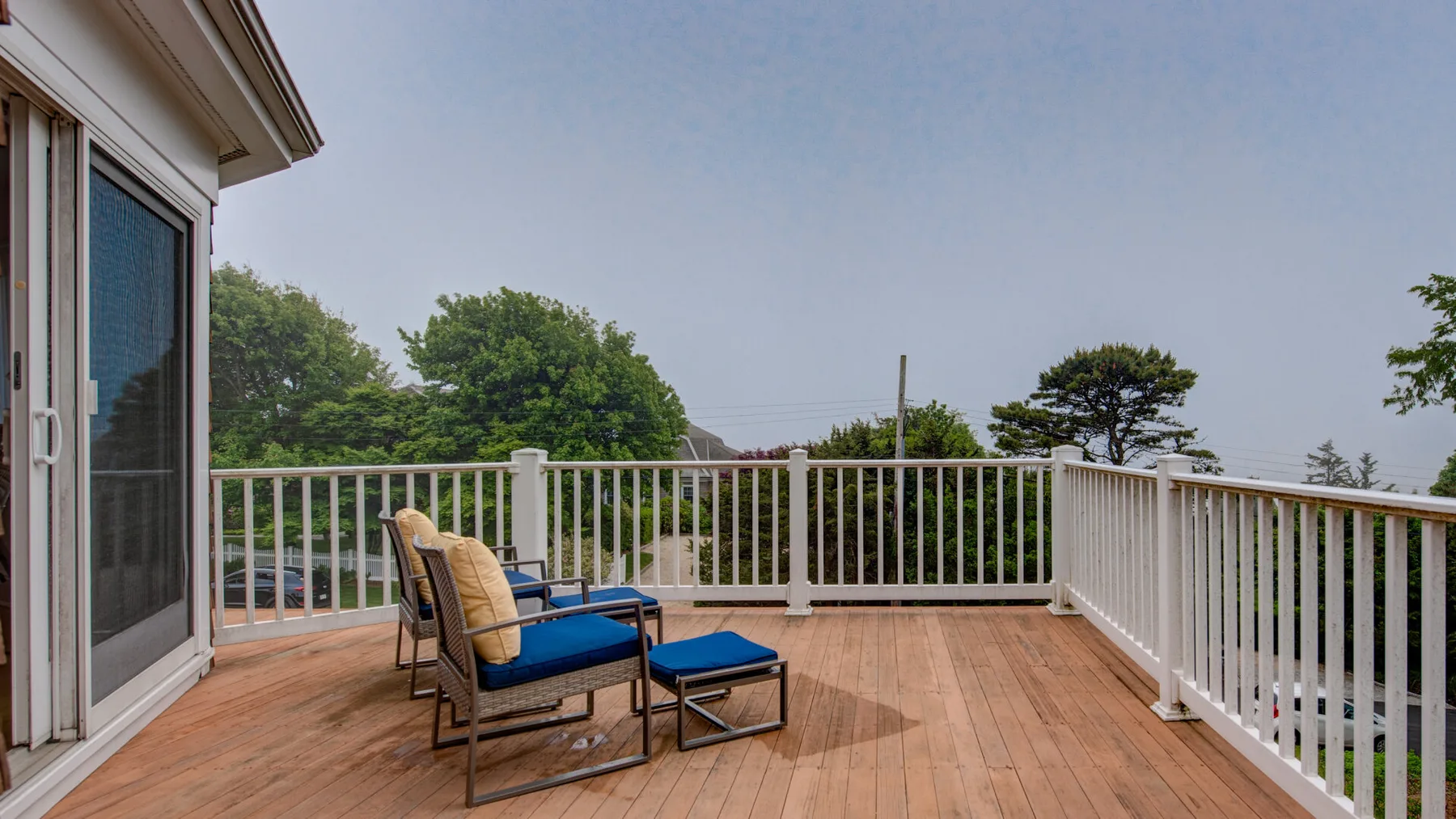 A wooden deck with outdoor seating overlooks trees beyond a white railing.