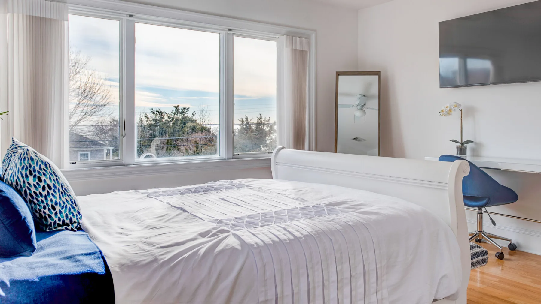 A bright bedroom has a white bed beside large windows and a blue chair.
