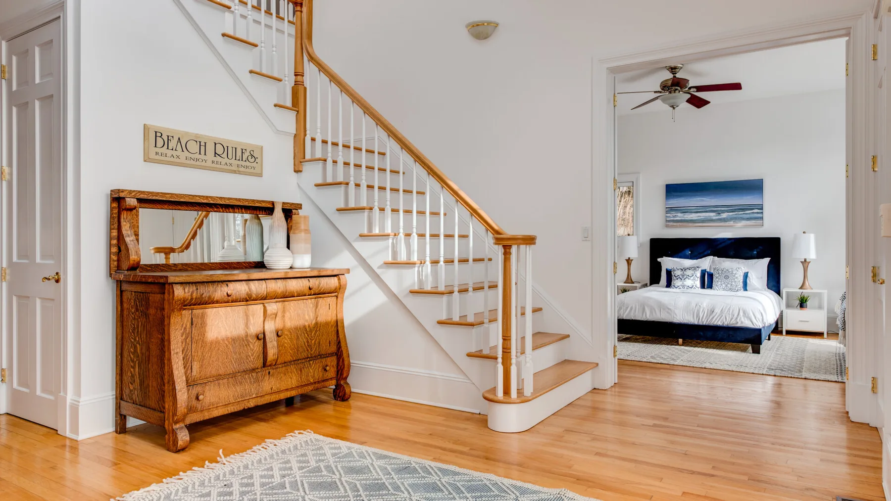 A bright hallway with a wooden staircase and a bedroom visible through the doorway.