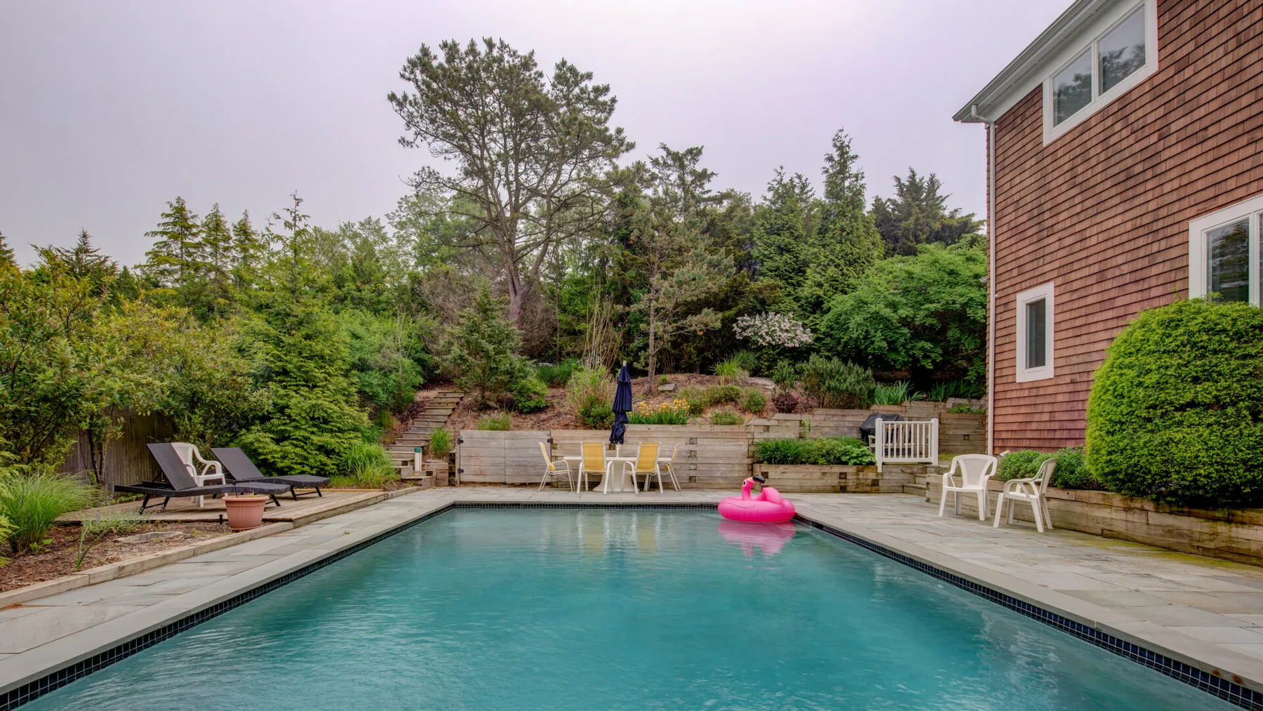 Una piscina al aire libre junto a una terraza, con jardines y un edificio cercano.