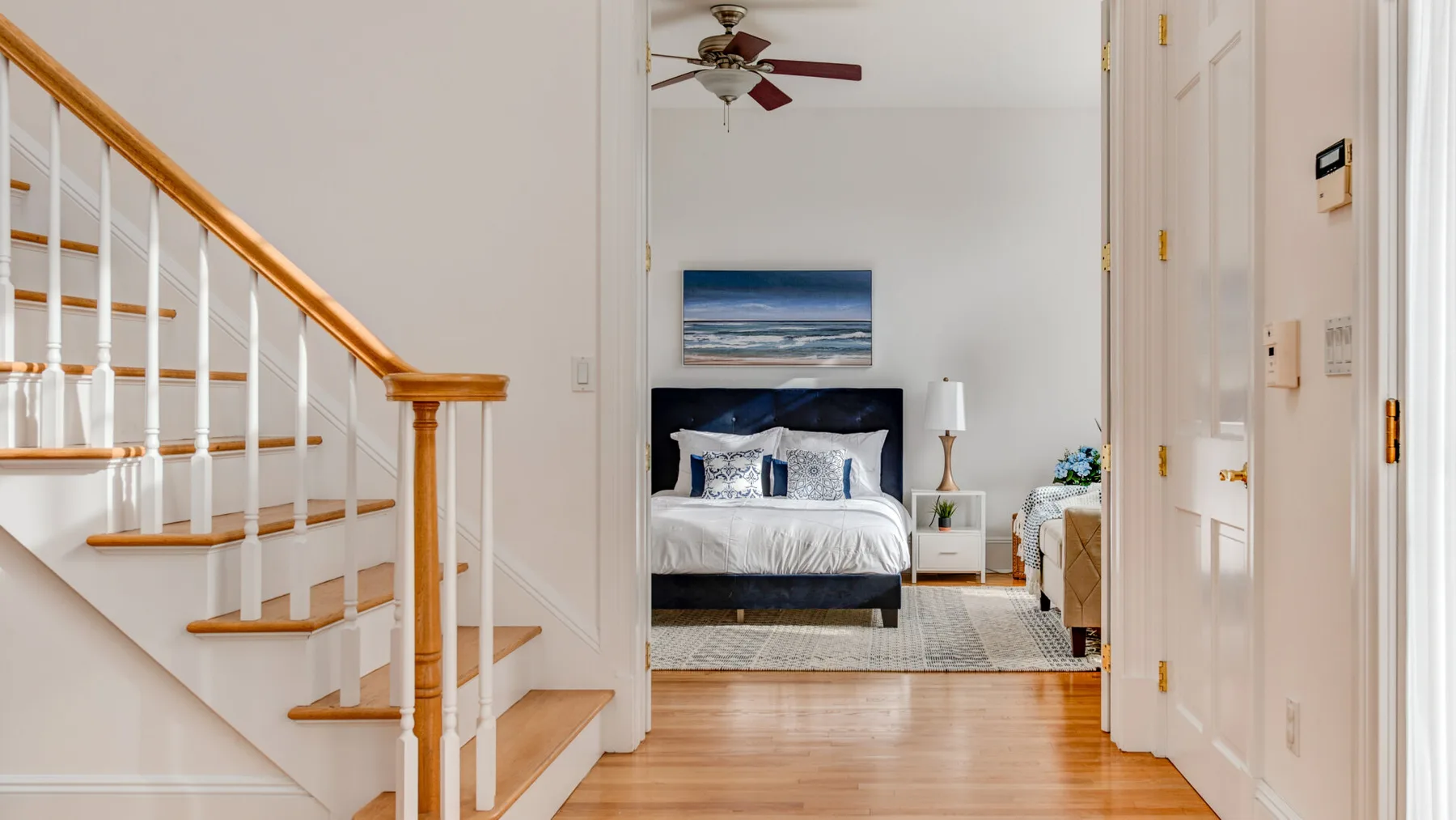 A hallway with a staircase opens toward a bedroom with a bed and ocean-themed artwork.