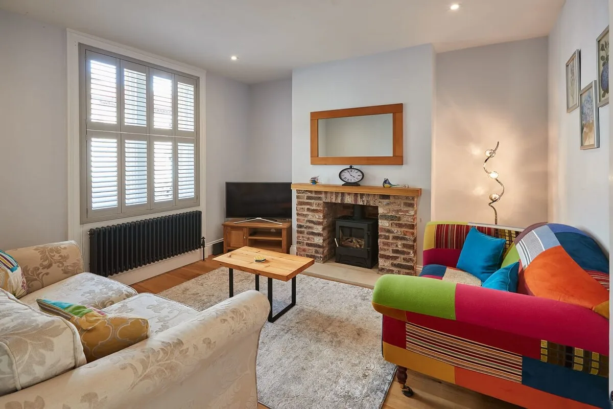 A bright living room with seating, a brick fireplace, and a large shuttered window.