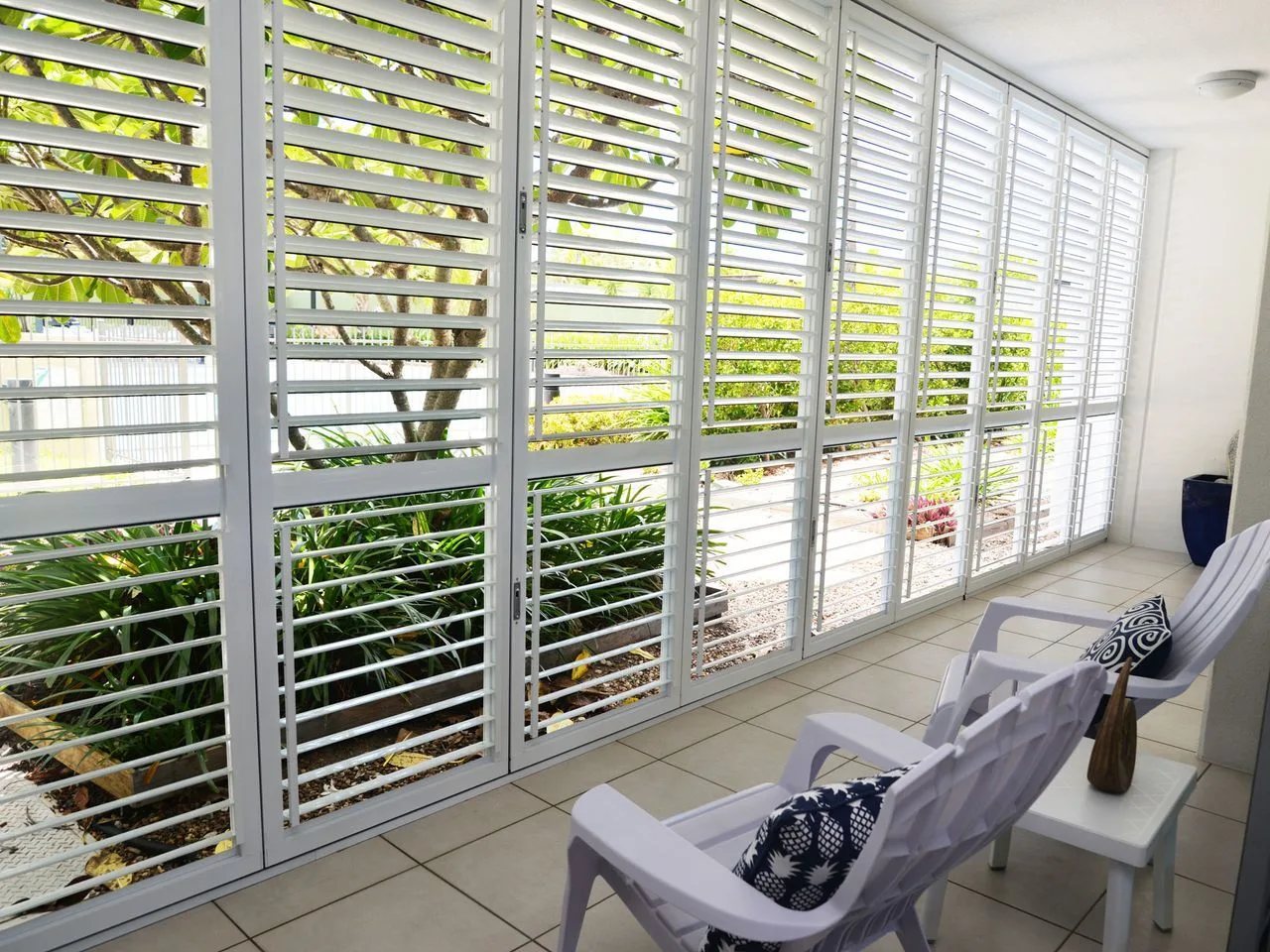 A bright enclosed patio with white seating beside large slatted windows.