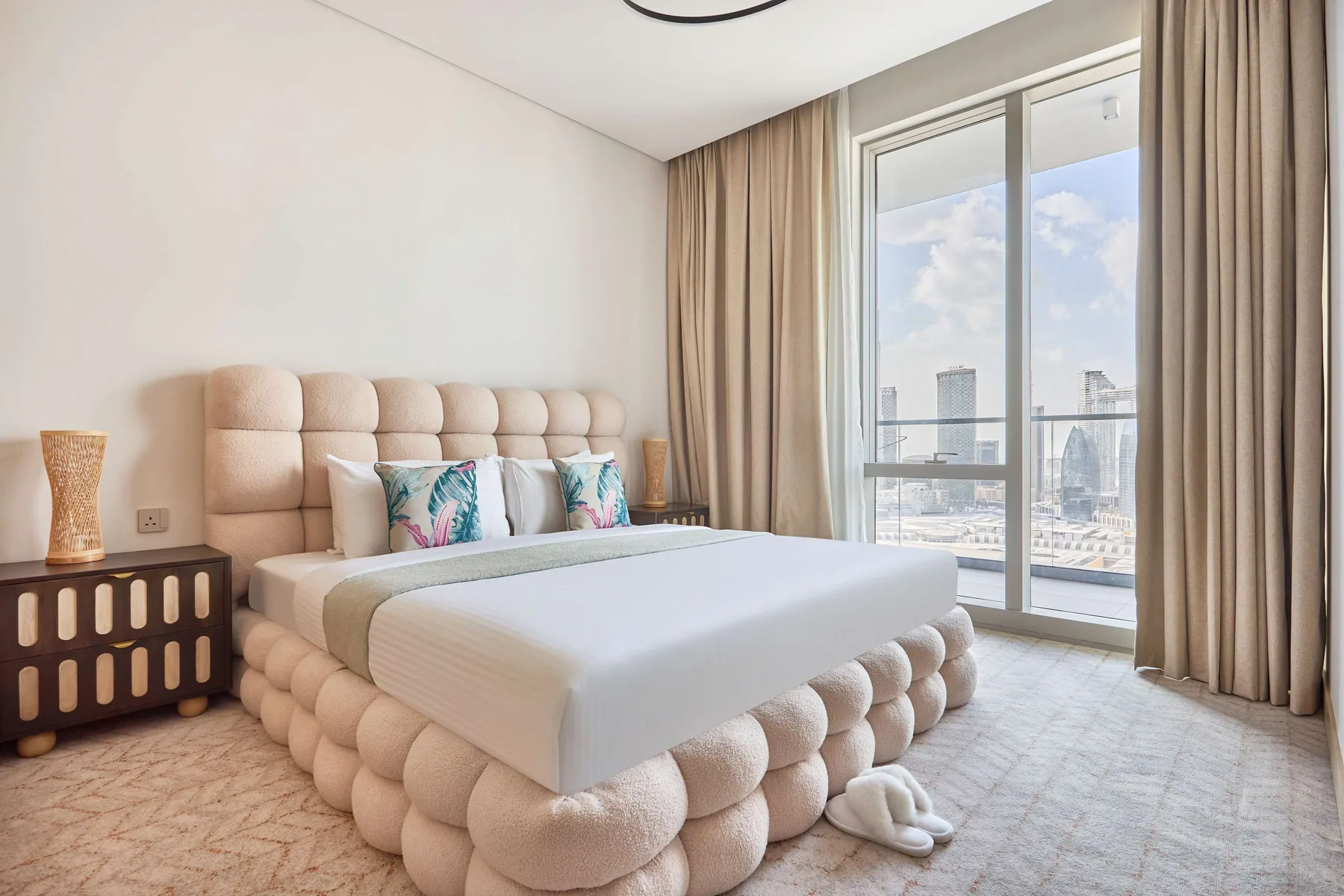 A bedroom with a tufted bed beside a large window overlooking the city.